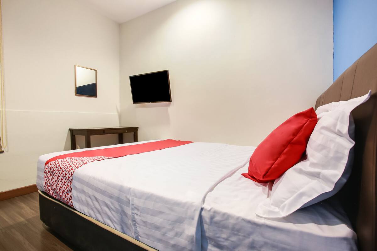 Ein ordentlich bezogenes Bett mit einem roten Kopfkissen und weißen Bettwäsche, ein kleiner Tisch, ein Flachbildfernseher und ein Spiegel an der Wand.
