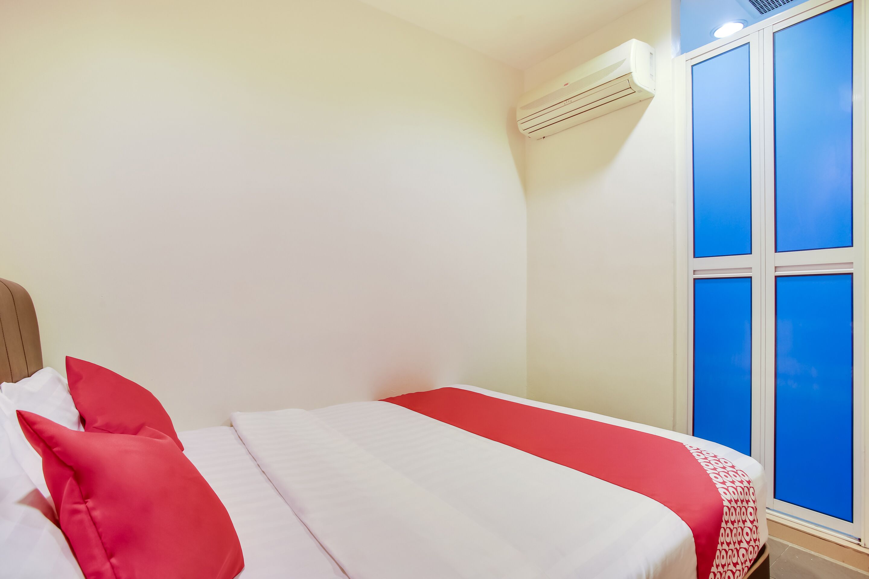 A hotel room with a bed, red and white bedding, a window with blue shutters, and an air conditioning unit.