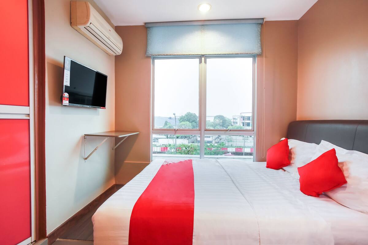 Ein Hotelzimmer mit Bett, roten Kissen, einem Fernseher an der Wand und einem großen Fenster.