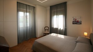 A bedroom with a bed, curtains, a chair, a wall-mounted lamp, and a painting.
