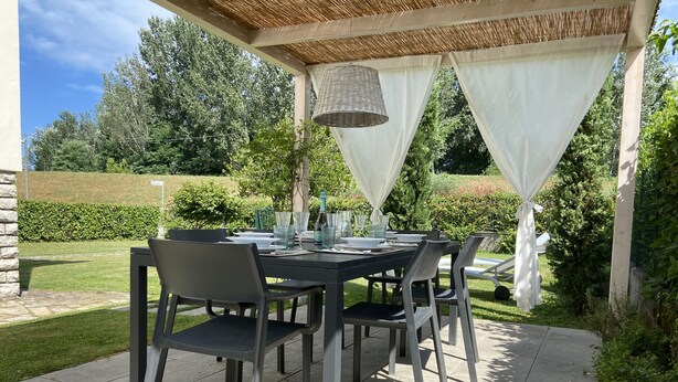 An outdoor patio with a table set for four, surrounded by chairs, under a pergola with white curtains.