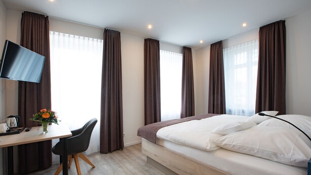 A modern hotel room with a bed, a desk with a chair, a TV, and a window with curtains. - Caesaro Hotel & Pizzeria (Hagen am Teuteburger Wald)