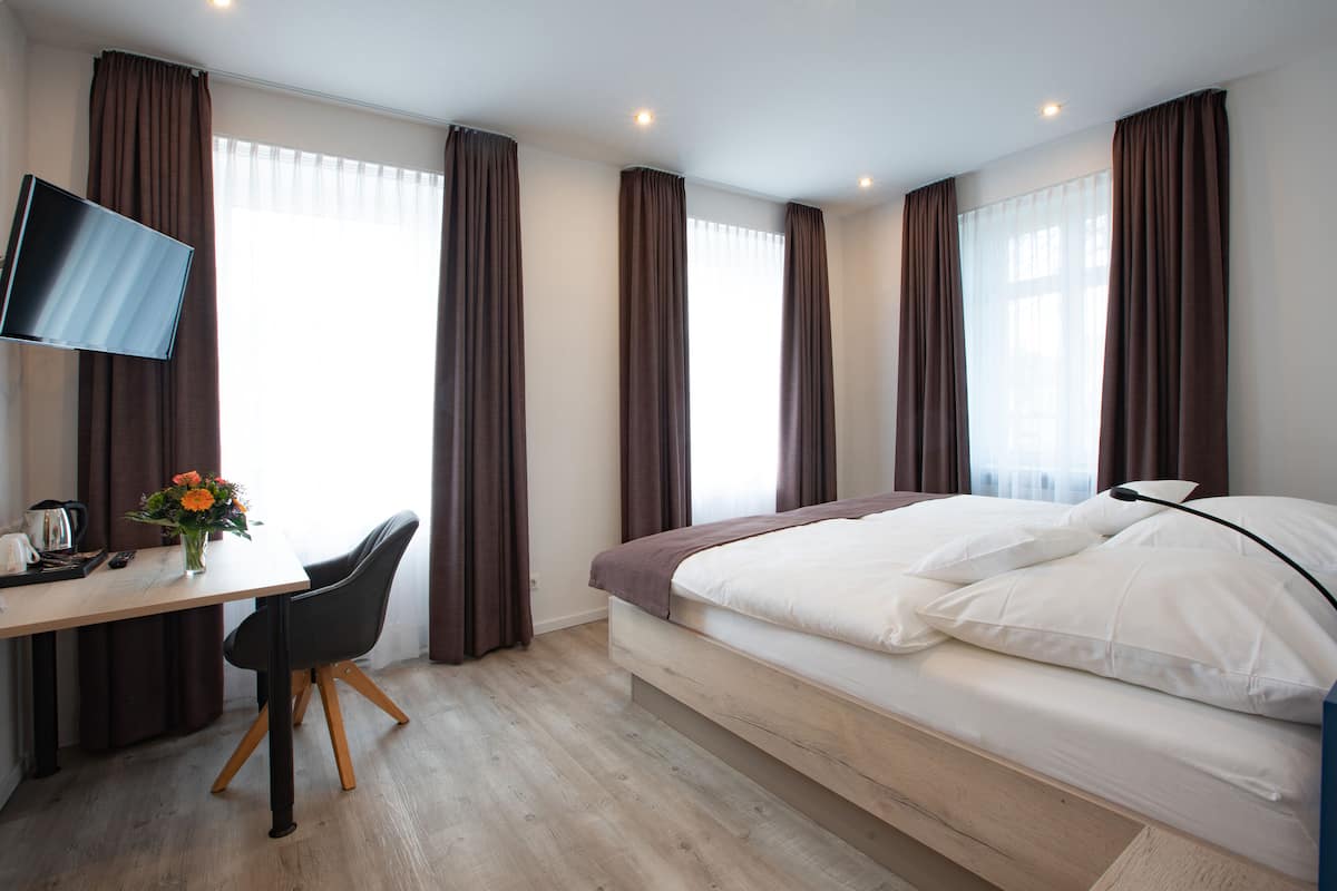 A modern hotel room with a bed, a desk with a chair, a TV, and a window with curtains.