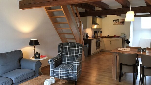 Een gezellige woonkamer met een geruite fauteuil, een grijze bank en een houten salontafel. Achtergrond toont een keuken met een eethoek.