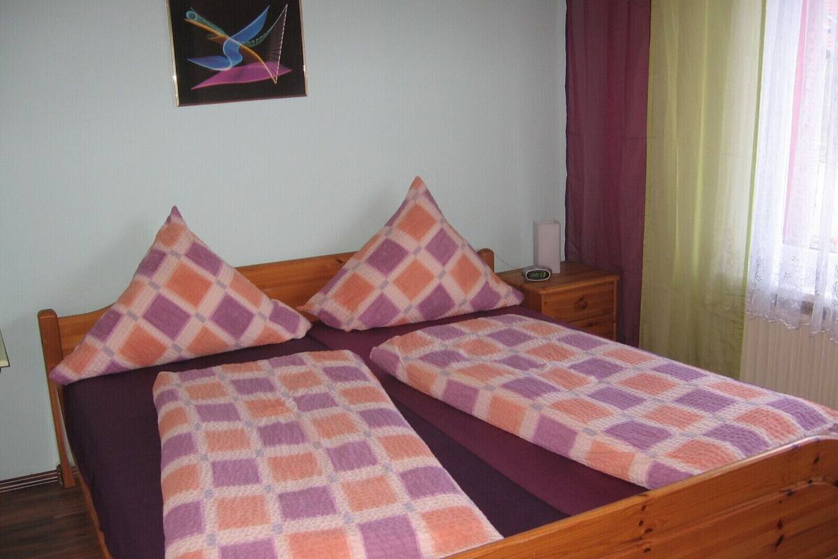 A bed with a checkered bedspread and two pillows, a wooden headboard, a nightstand with a lamp, and a painting on the wall.