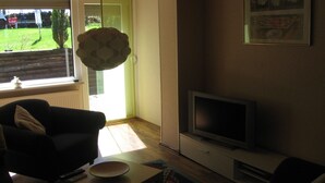 A living room with a sofa, a television, and a window overlooking a garden. - Ferienwohnungen an der Loipe (Braunlage)