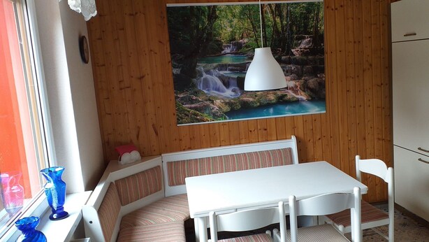 A small dining area with a white table and bench, a hanging pendant light, and a kitchen area with cabinets. - Ferienwohnungen an der Loipe (Braunlage)