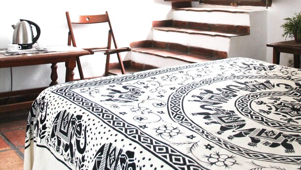 Une chambre avec un lit recouvert d’une couvre-lit à motifs, une chaise en bois, une table avec une bouilloire et une fenêtre donnant sur un paysage verdoyant.
