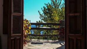Une porte en bois ouverte donne sur un balcon doté d’un sol en pierre, d’une rambarde en métal et d’une vue sur des arbres et un ciel bleu.