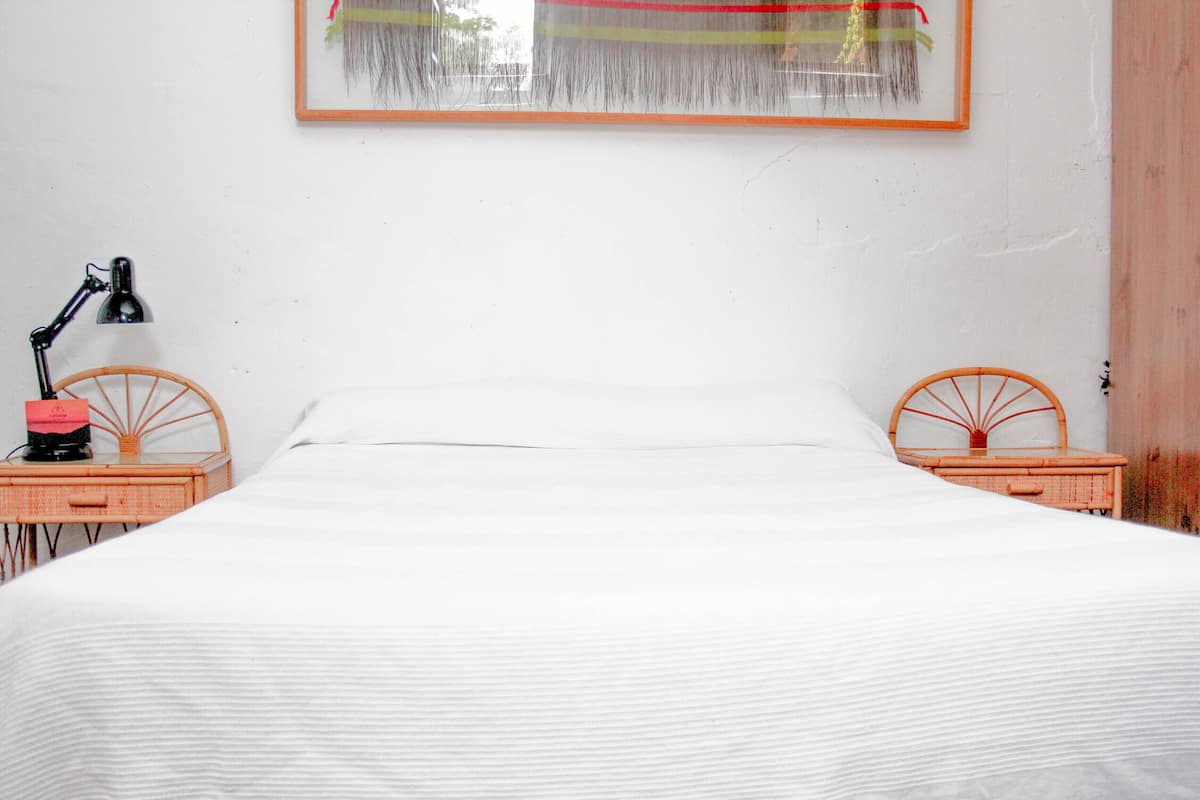 A bed with white bedding, two wicker nightstands with lamps, and a framed artwork above the bed.