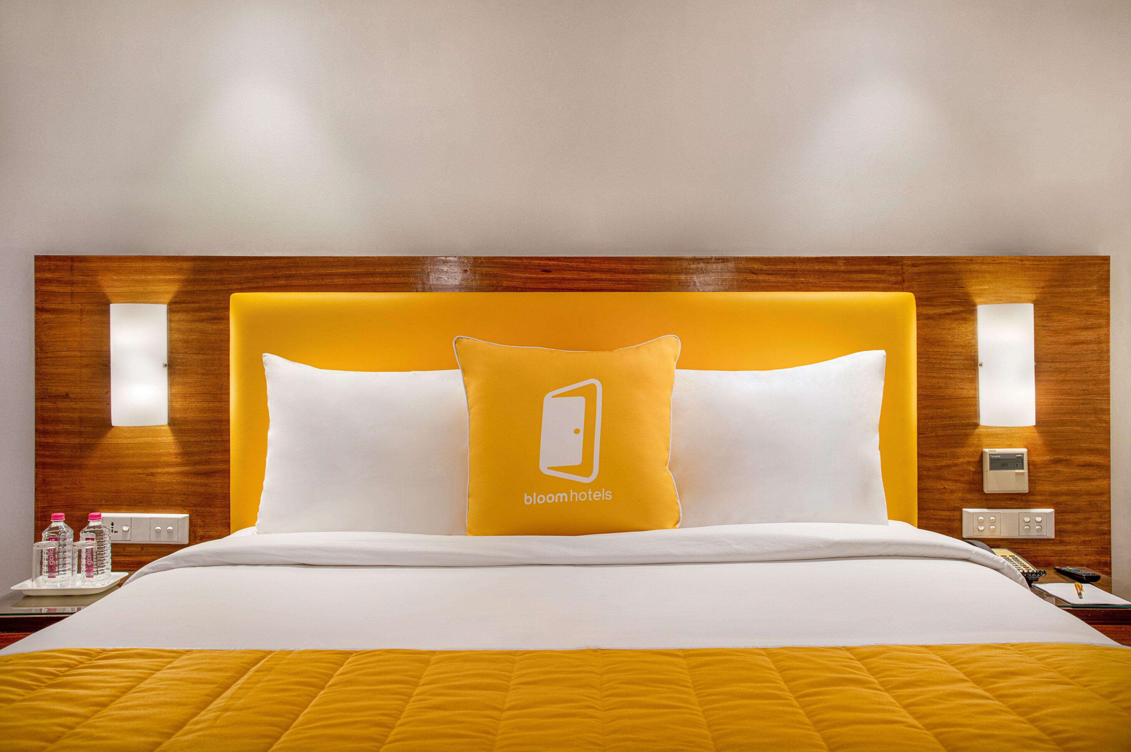 A hotel room with a wooden headboard, a bed with white and yellow bedding, a single yellow pillow with a logo, bedside tables with water bottles, and a control panel on the wall.