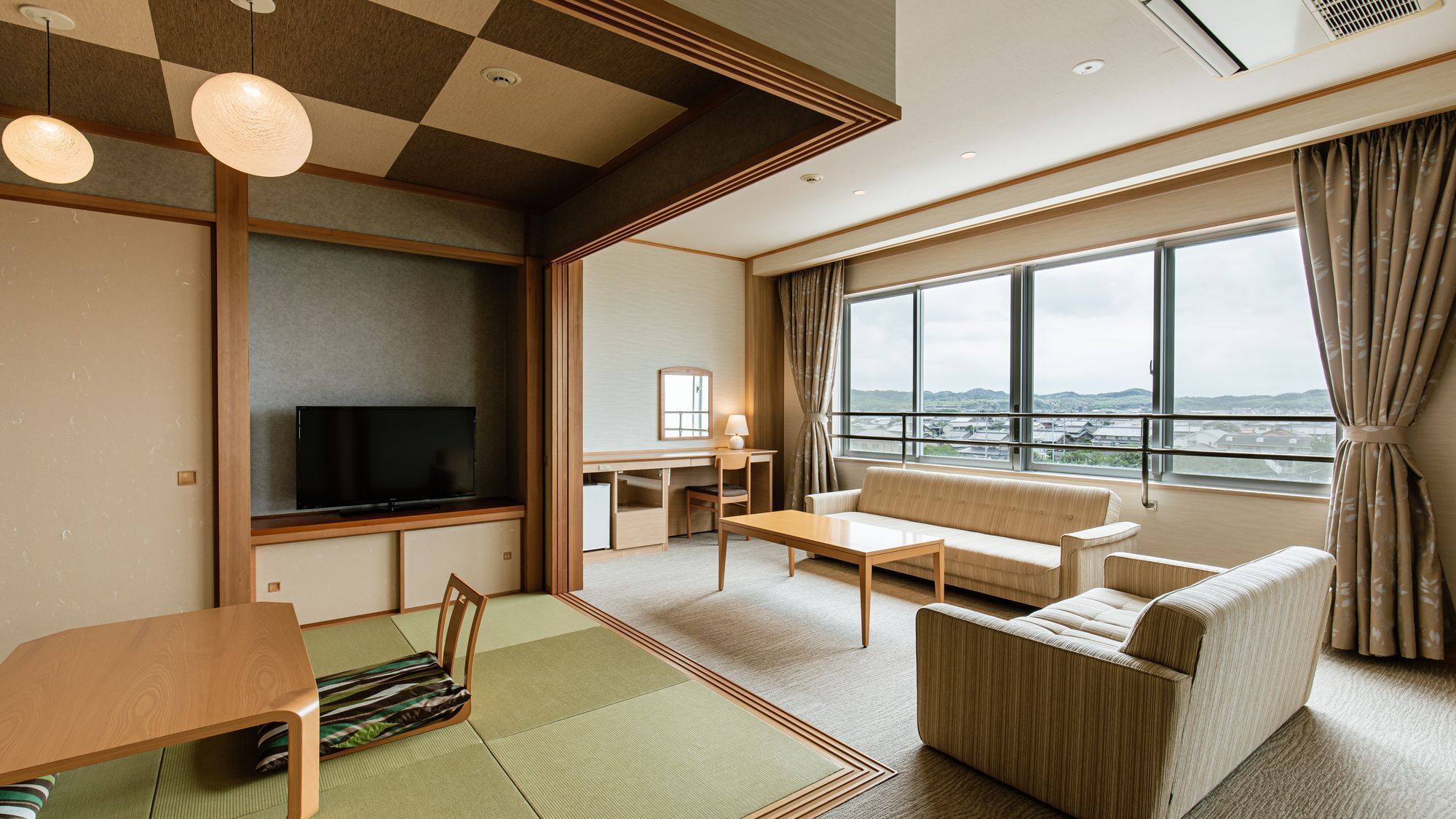 Habitación de hotel moderna con televisor, sofá, mesa de comedor y ventanas amplias con vistas a la ciudad.