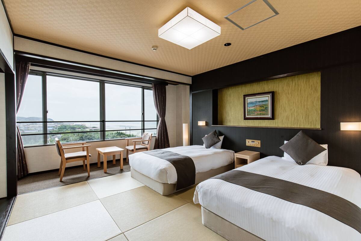 A hotel room with two beds, a desk, and a chair. There is a large window providing a view of the outdoors.