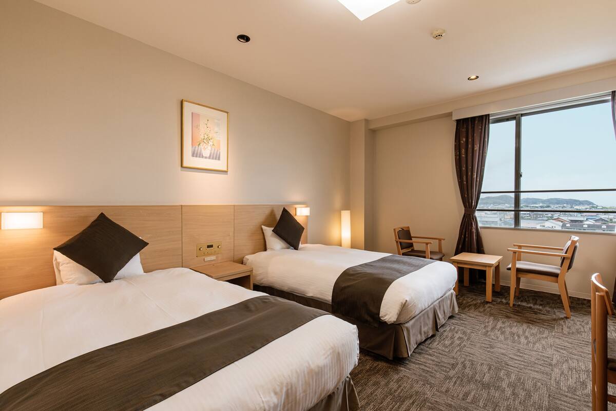 A hotel room with two beds, a desk, and a chair. There is a window with a view of buildings.