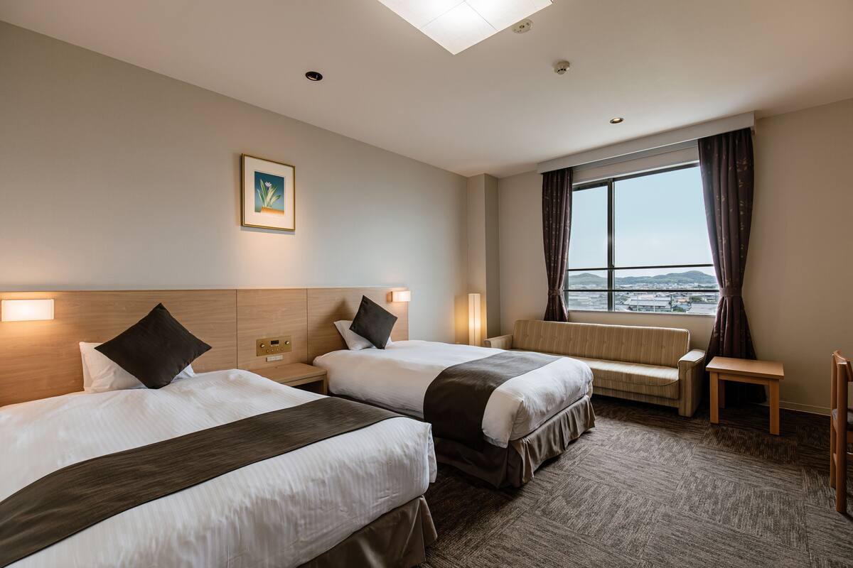 A hotel room with two beds, a sofa, a small table, and a large window with a view.
