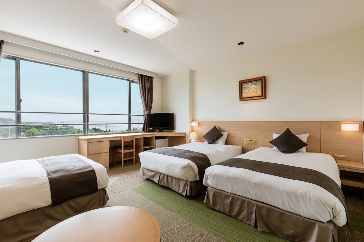 A hotel room with two beds, a desk, a television, and a large window offering a view of the outdoors.