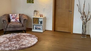 Een woonkamer met een bruine leren fauteuil, een rond tapijt, een witte plank met decoratieve objecten, een houten deur en een wandlamp.