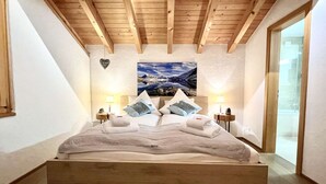 Een slaapkamer met een houten plafond, een groot bed met kussens, twee nachtkastjes met lampen en een schilderij van een berglandschap aan de muur.