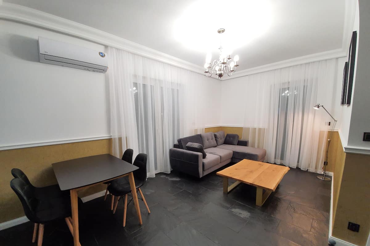 A modern living room with a sofa, a wooden coffee table, a dining table with chairs, and a chandelier.