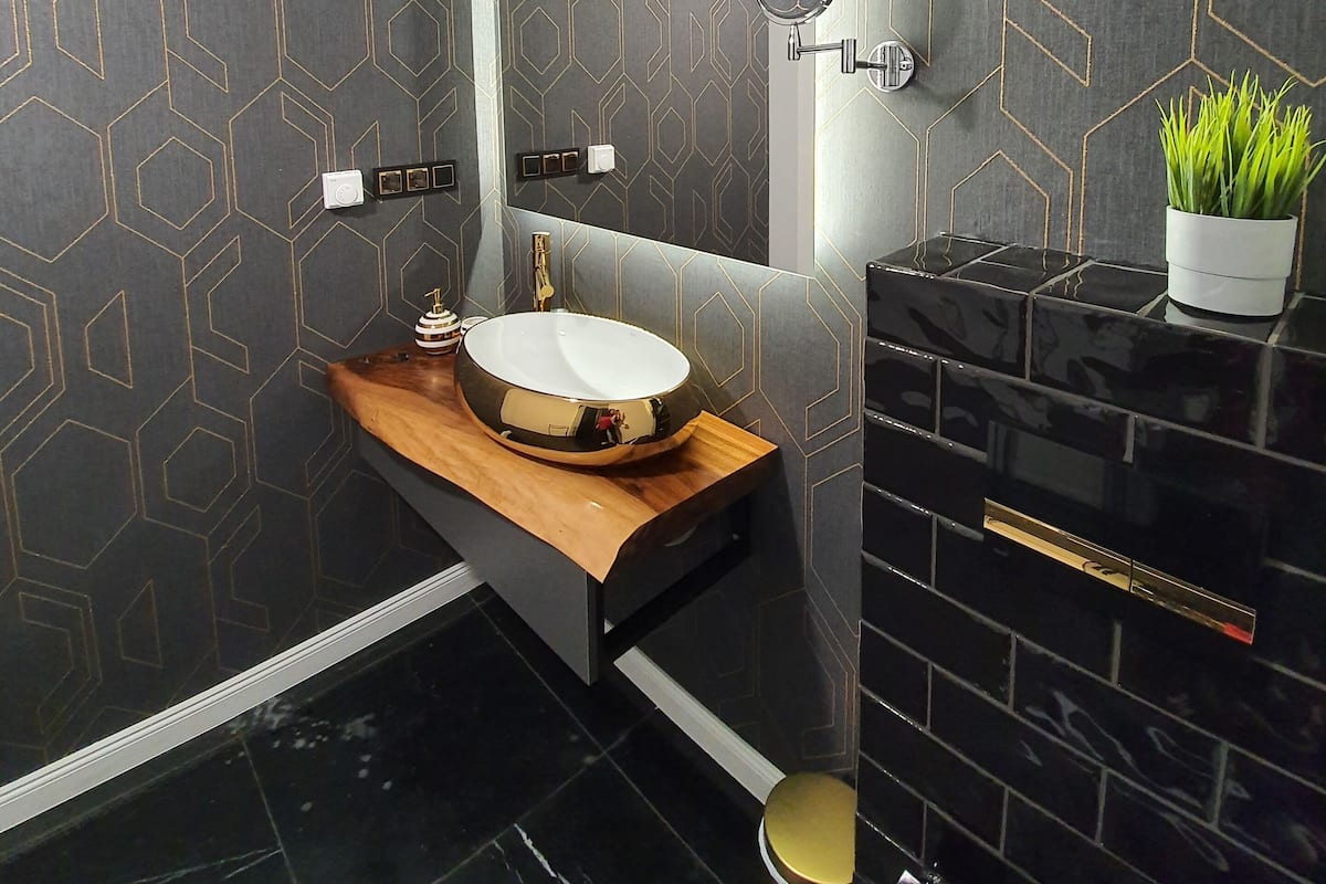 A modern bathroom with a geometric patterned wall, a wooden vanity, a round sink, a black tiled shower area, and a potted plant.