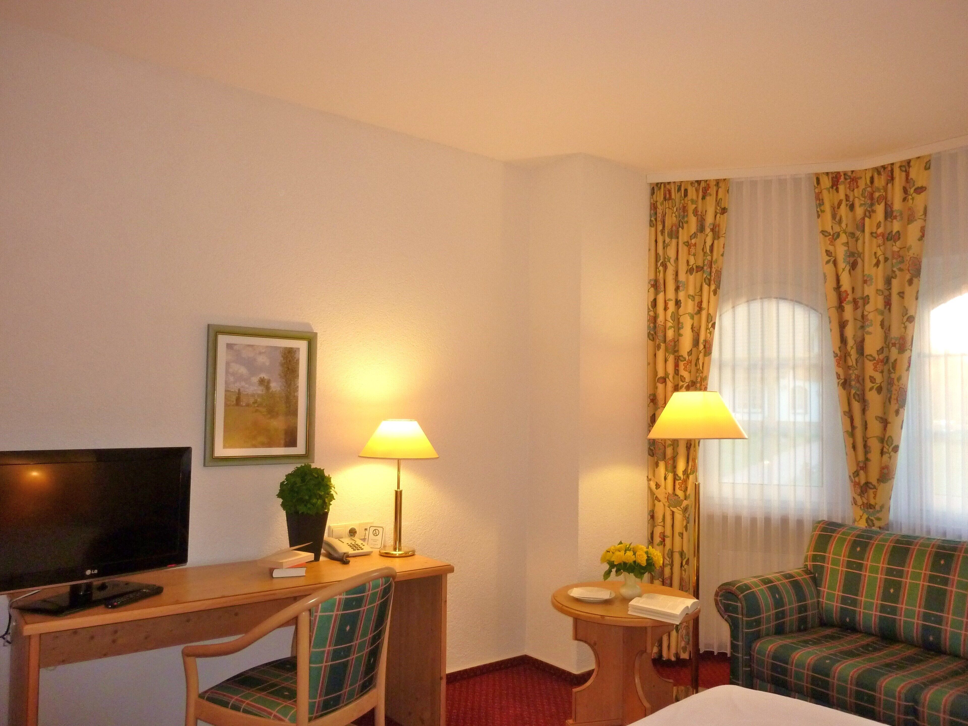 Habitación de hotel con una cama, un escritorio con televisor, dos lámparas, una silla, un sofá y una mesita con un jarrón de flores.