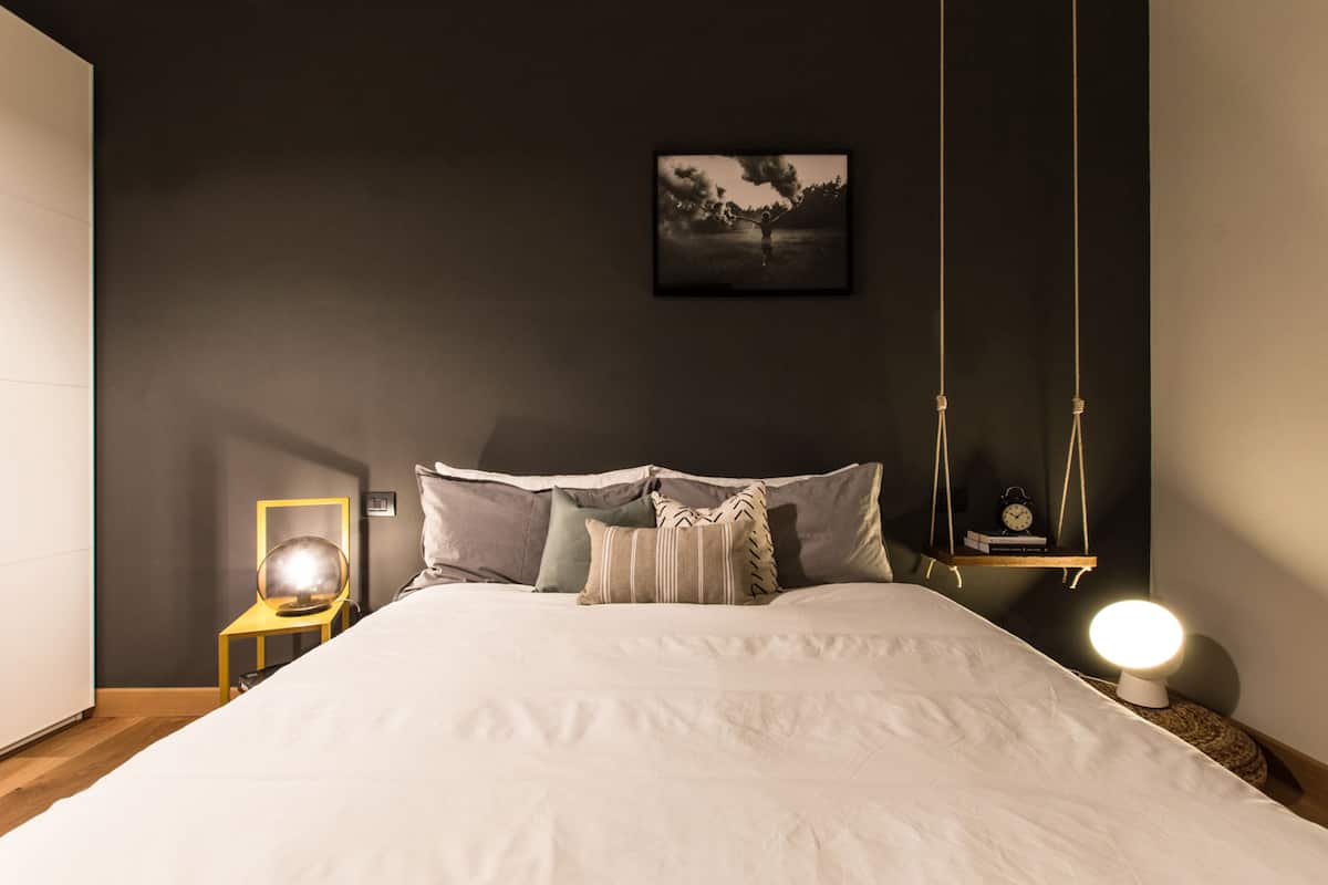 A bedroom with a large bed, a bedside table with a lamp, a wall-mounted clock, and a framed picture on the wall.