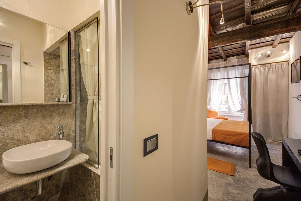 A bathroom with a marble sink, a glass shower enclosure, and a bed with an orange bedspread.