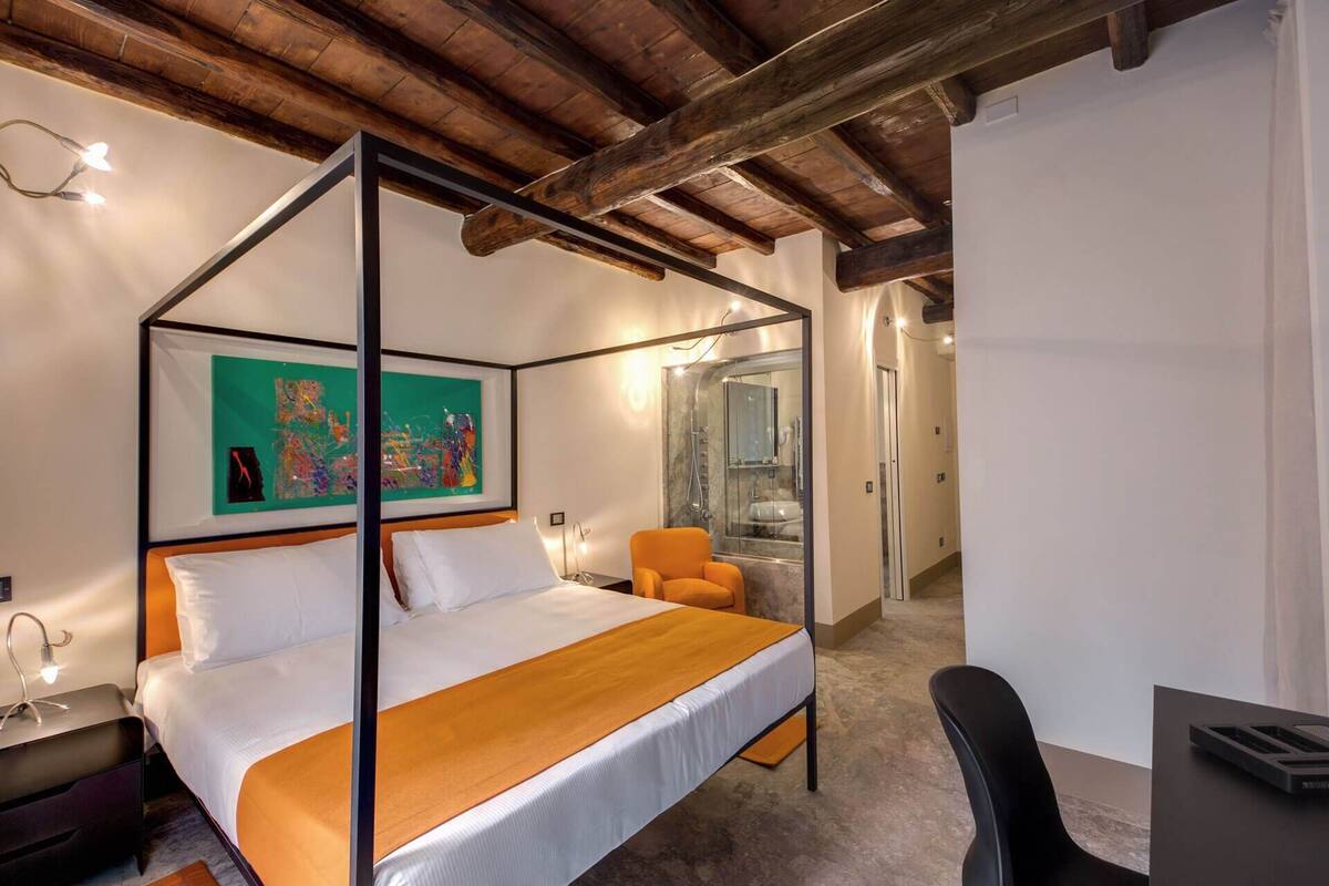 A modern bedroom with a four-poster bed, a small desk, a chair, and a glass-enclosed shower.