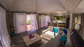 A spacious living area with a stone wall, wooden ceiling, and a stone fireplace. There is a sofa with colorful cushions and a wooden coffee table with flowers. - Cabañas Caballieri (Paiguano)