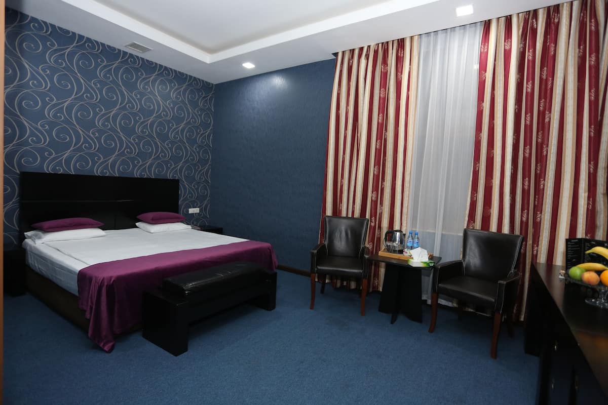 A hotel room with a bed, two chairs, a small table with a kettle and a fruit bowl, and a window with striped curtains.