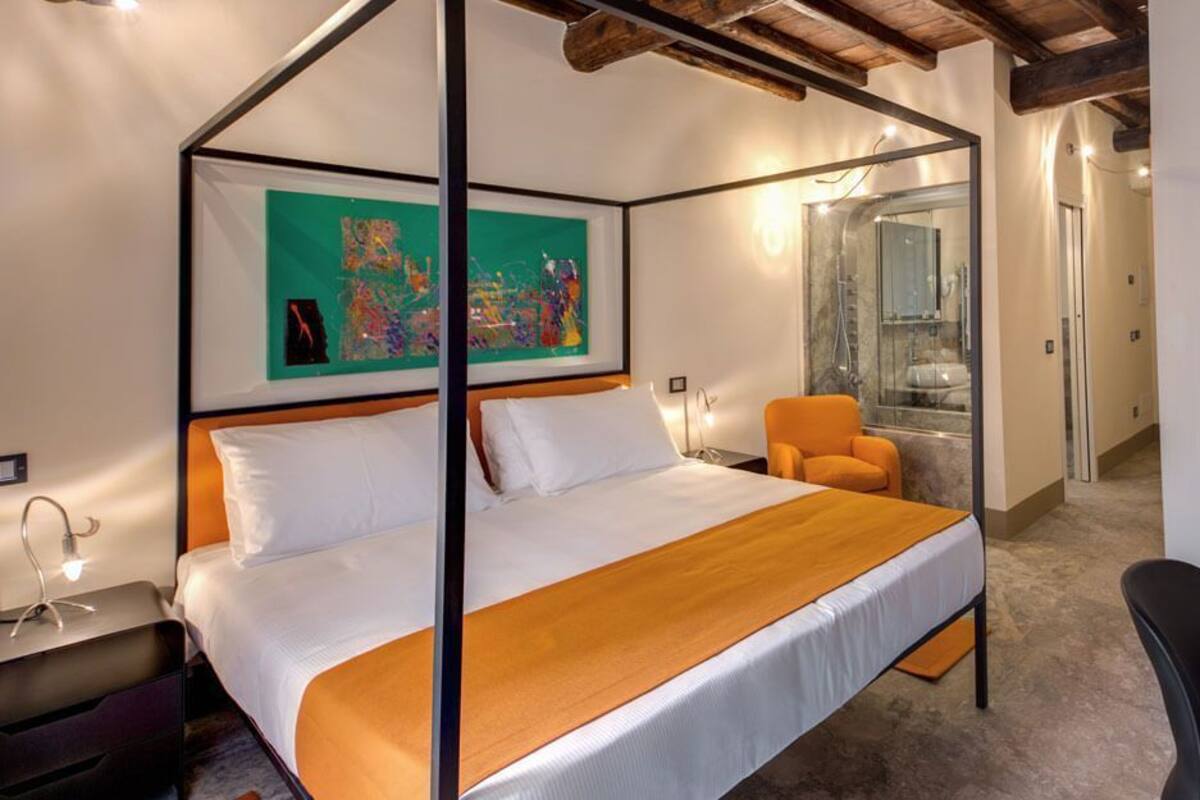 A four-poster bed with a wooden headboard, a single orange chair, a desk with a black chair, and a bathroom with a glass shower.