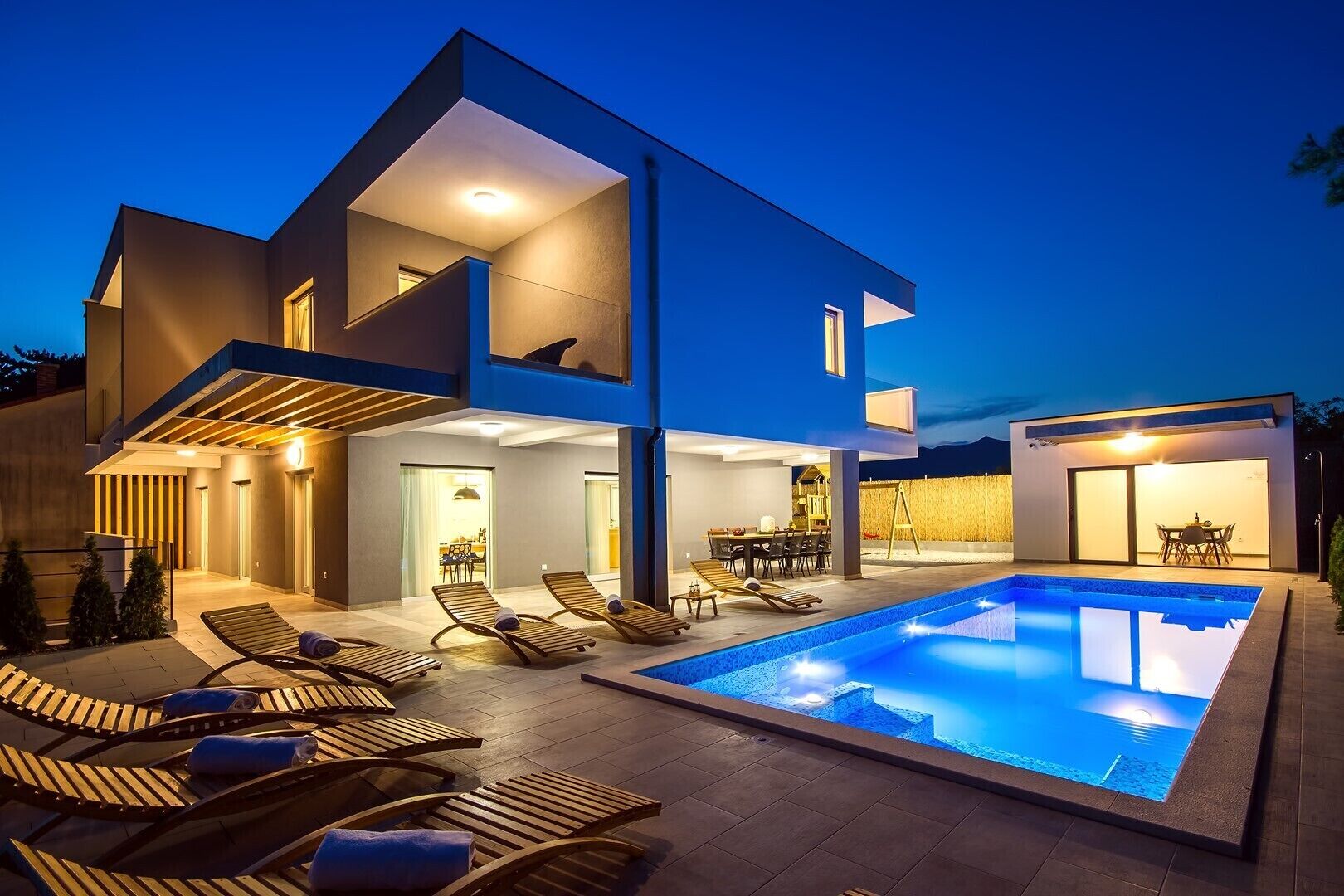 Una casa moderna de dos pisos con piscina, mobiliario exterior y un patio bien iluminado.