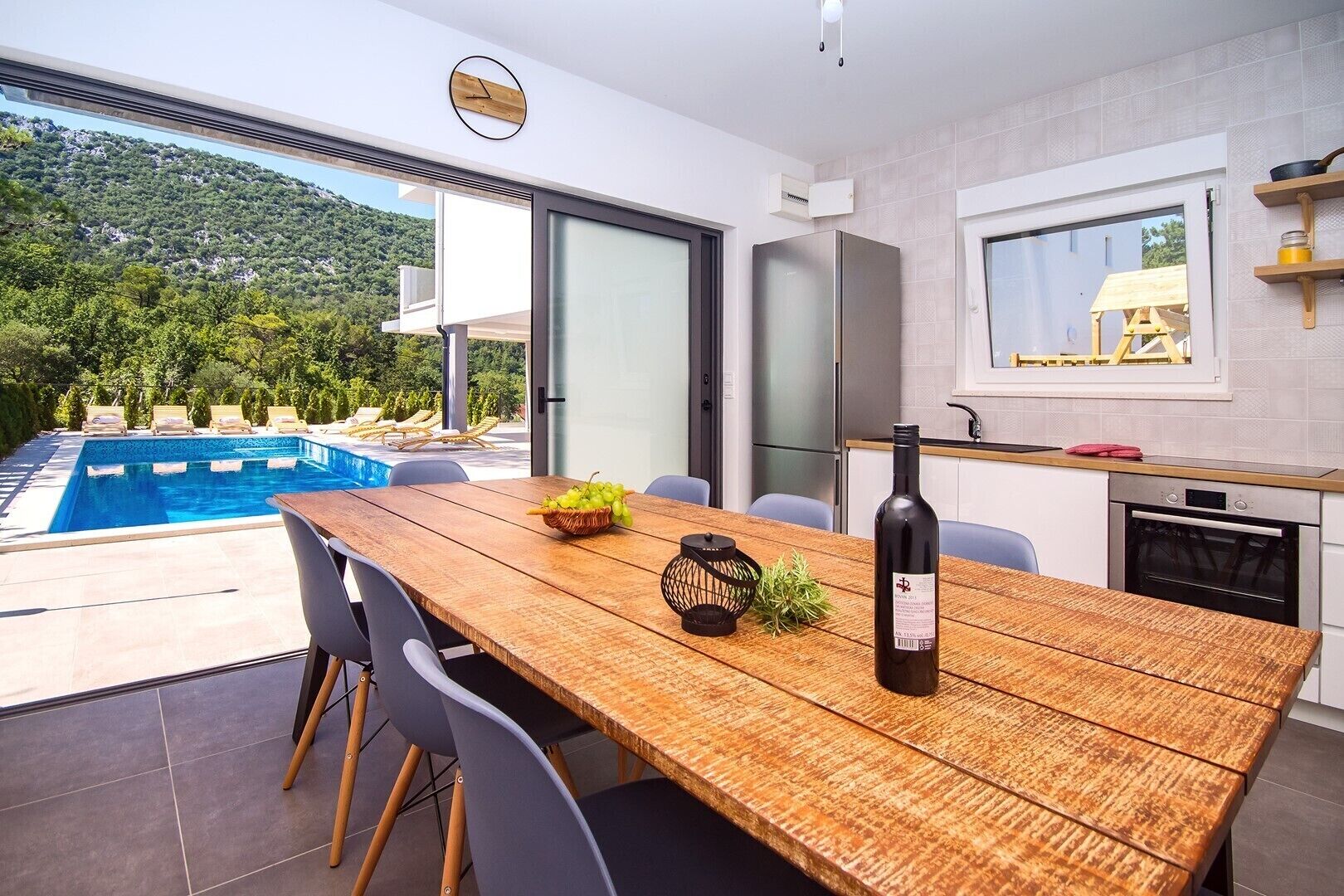 Une cuisine moderne dotée d’un four encastré, un coin repas avec une table et des chaises en bois, et une vue sur une piscine et un espace verdoyant.