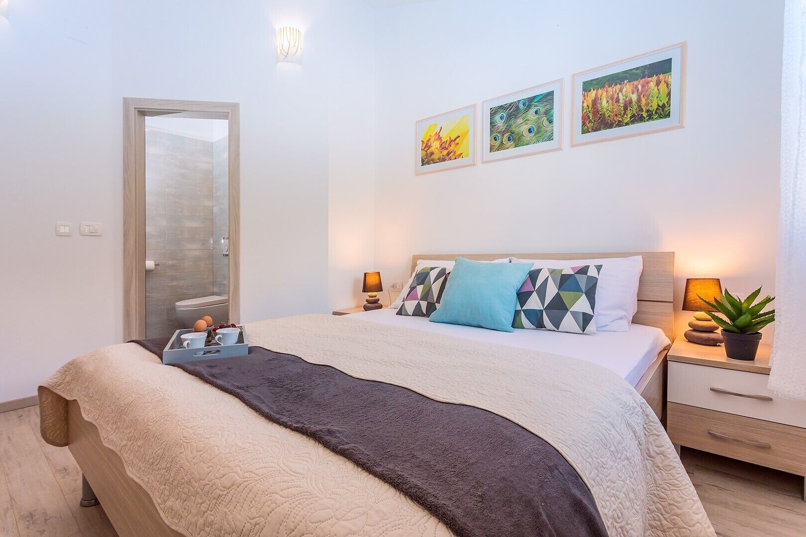 A modern bedroom with a large bed, a bedside table with a lamp, a small potted plant, and a mirror.