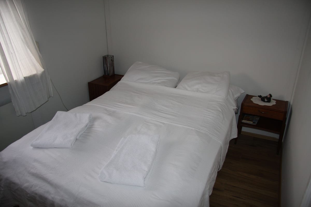 Cama bem arrumada com travesseiros e toalha dobrada, mesa de cabeceira com abajur e pequena mesa de apoio de madeira com um livro.