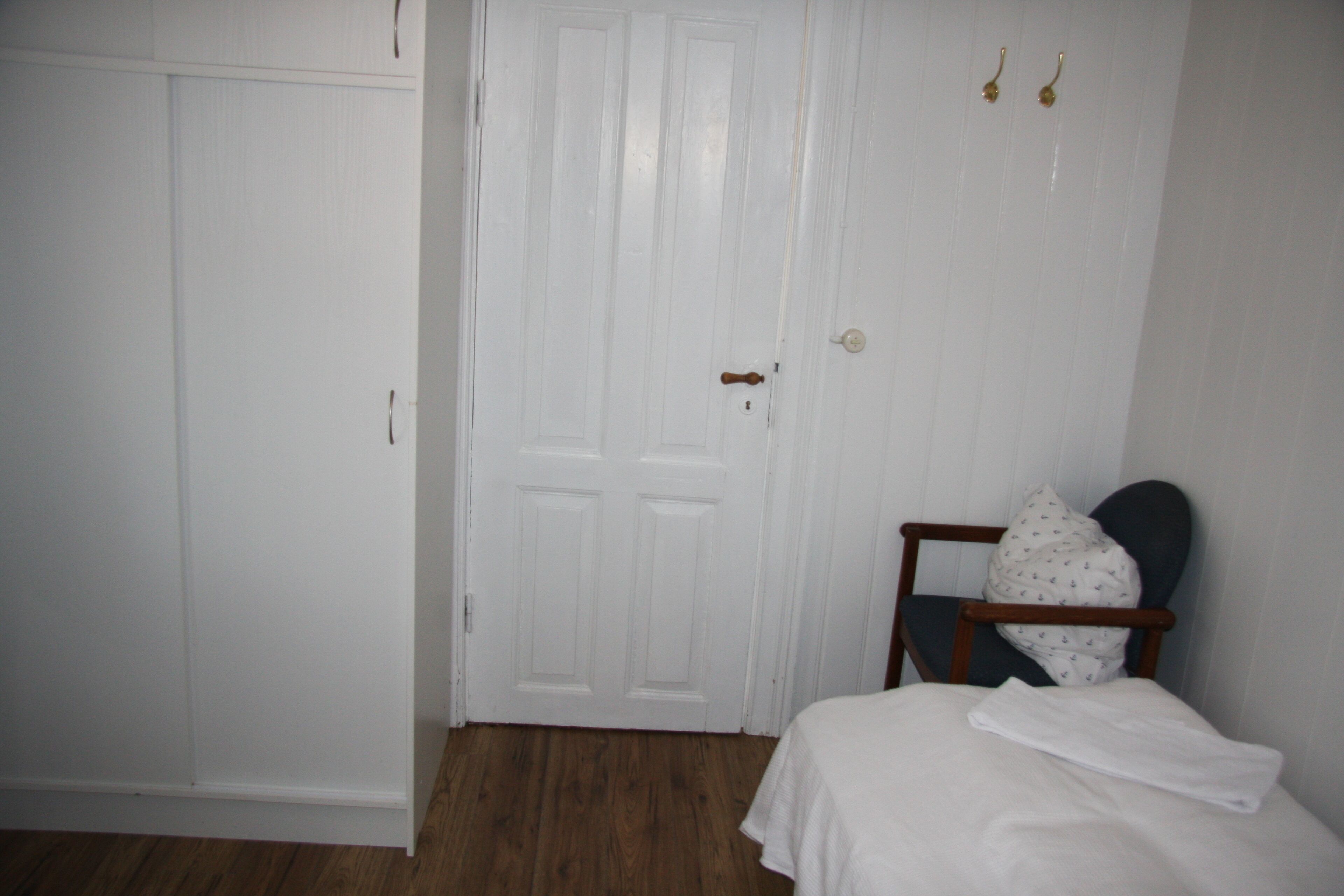 A bedroom with a wooden floor, a white bed with a white blanket, a wooden chair, and a white wardrobe.