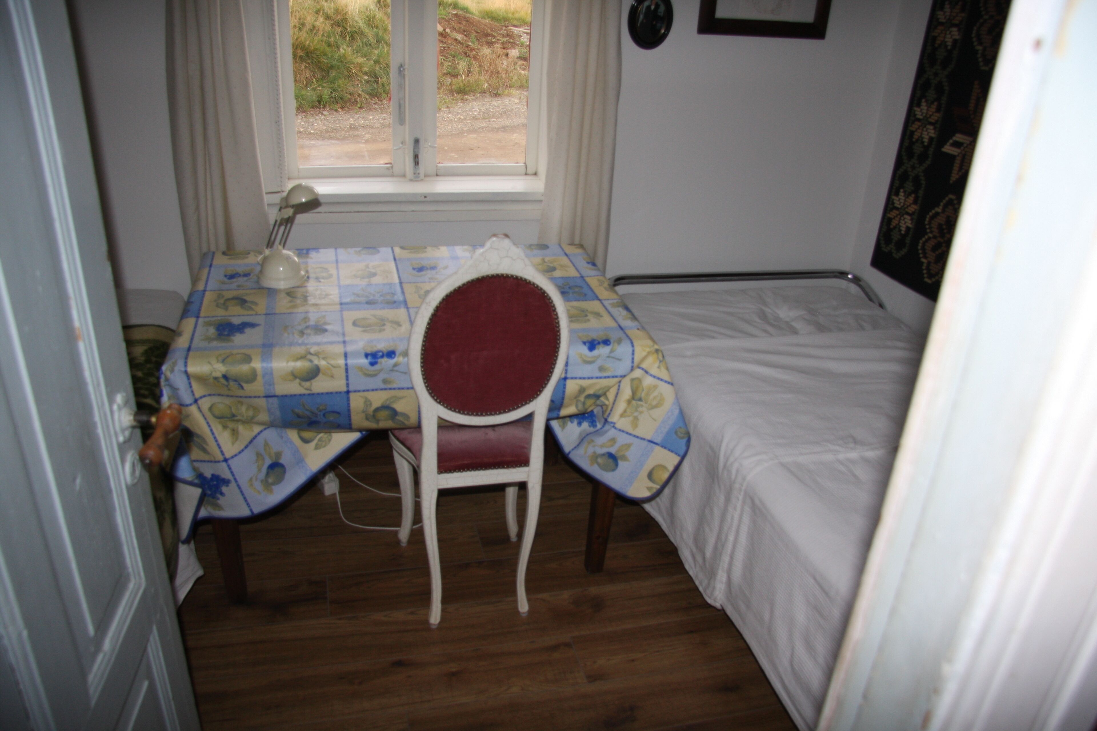 A small room with a bed, a table with a floral tablecloth, a chair, and a window with curtains.