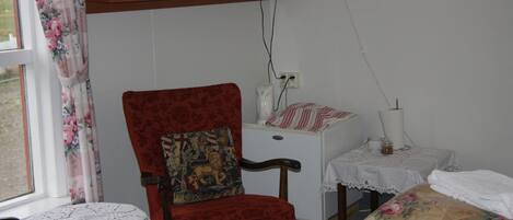 A room with a red upholstered chair, a small table with a lace tablecloth, a white bedside table with a lamp, and a television mounted on the wall.