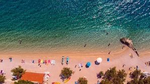 Vue aérienne d’une plage aux eaux turquoise et limpides, avec un promontoire rocheux, ainsi que plusieurs parasols et chaises longues.
