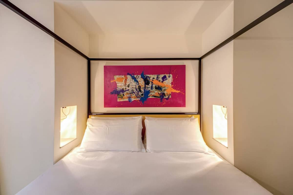 A modern hotel room with a large bed, white linens, and a colorful abstract painting above the headboard.