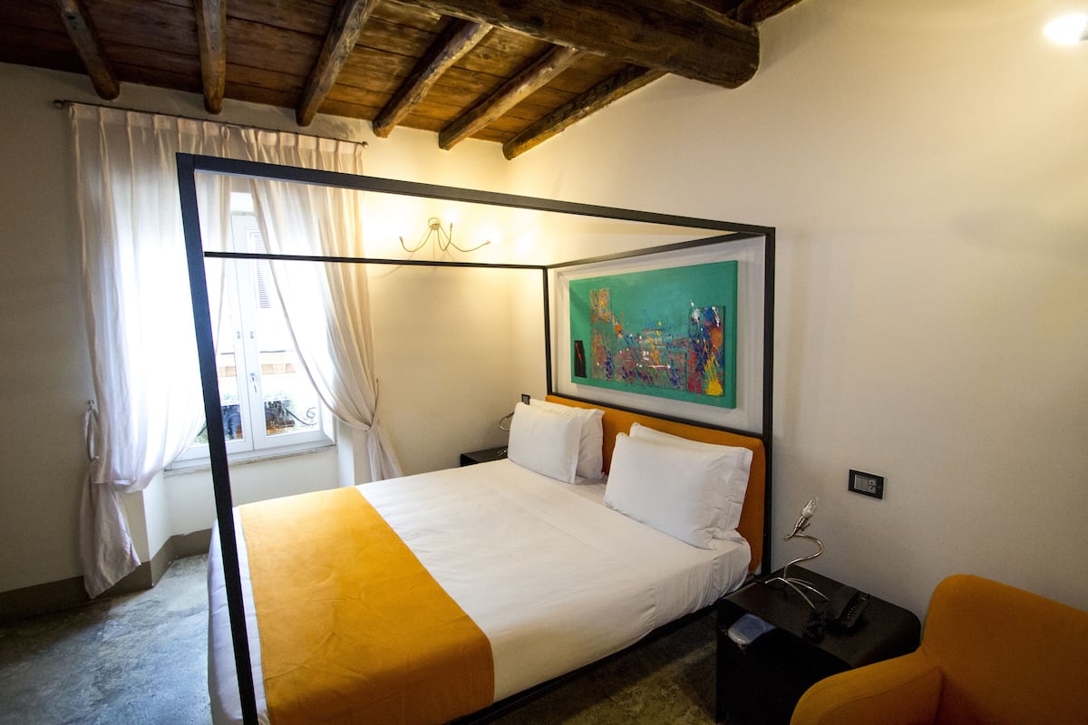 A four-poster bed with a white and yellow bedspread, a painting above the bed, and a window with curtains.