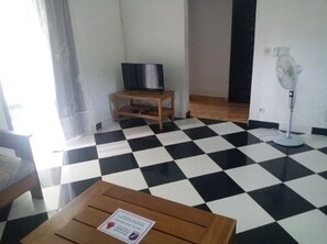 A room with a checkered floor, a wooden table with a sign, a television on a stand, and a standing fan.