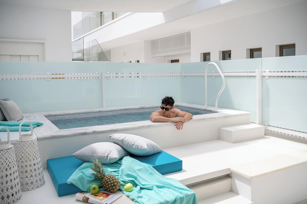 Una piscina interior moderna con una persona relajándose en el borde, rodeada de zonas de estar y una escalera.