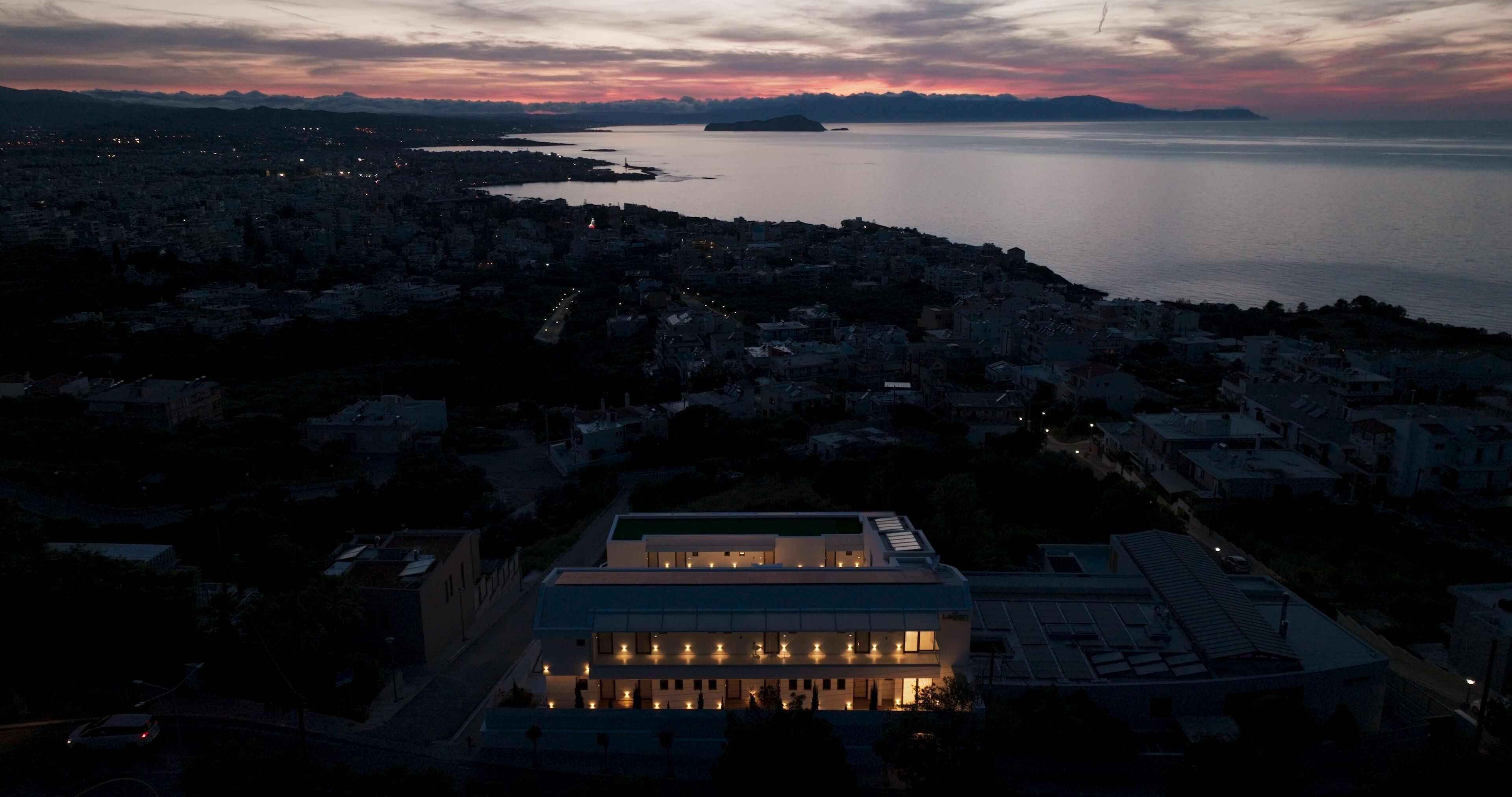 A coastal cityscape at dusk with a prominent modern building featuring multiple illuminated windows.