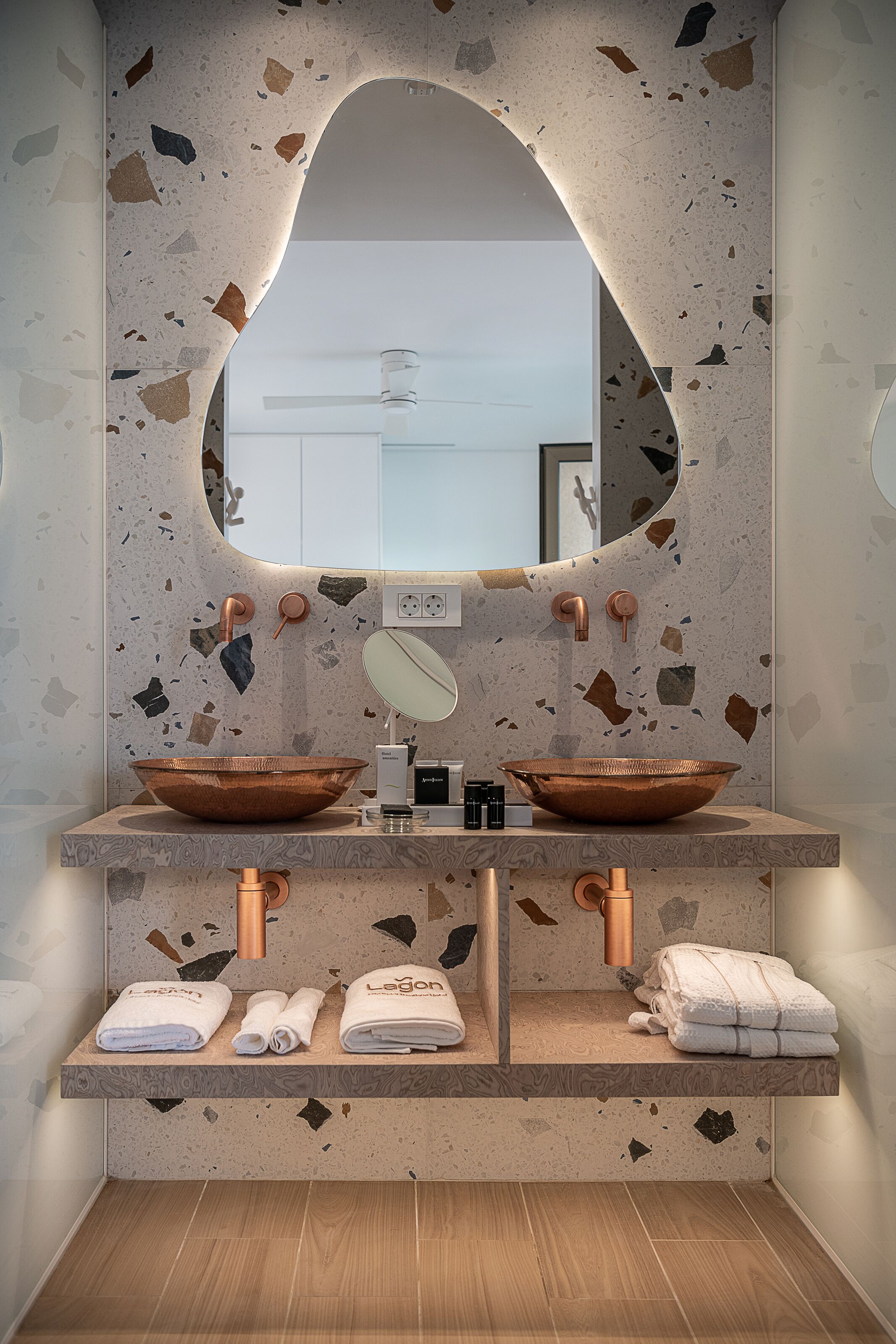 Et badeværelse med backsplash i terrazzo, to kobbervaske, et spejl og håndklæder på bordet.