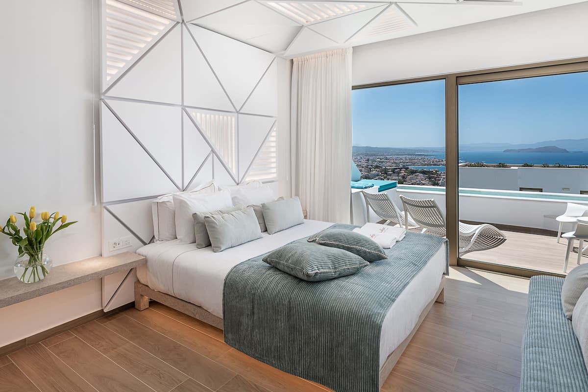 Un dormitorio moderno con una cama grande, vistas a la ciudad y al mar, y un balcón con mobiliario exterior.