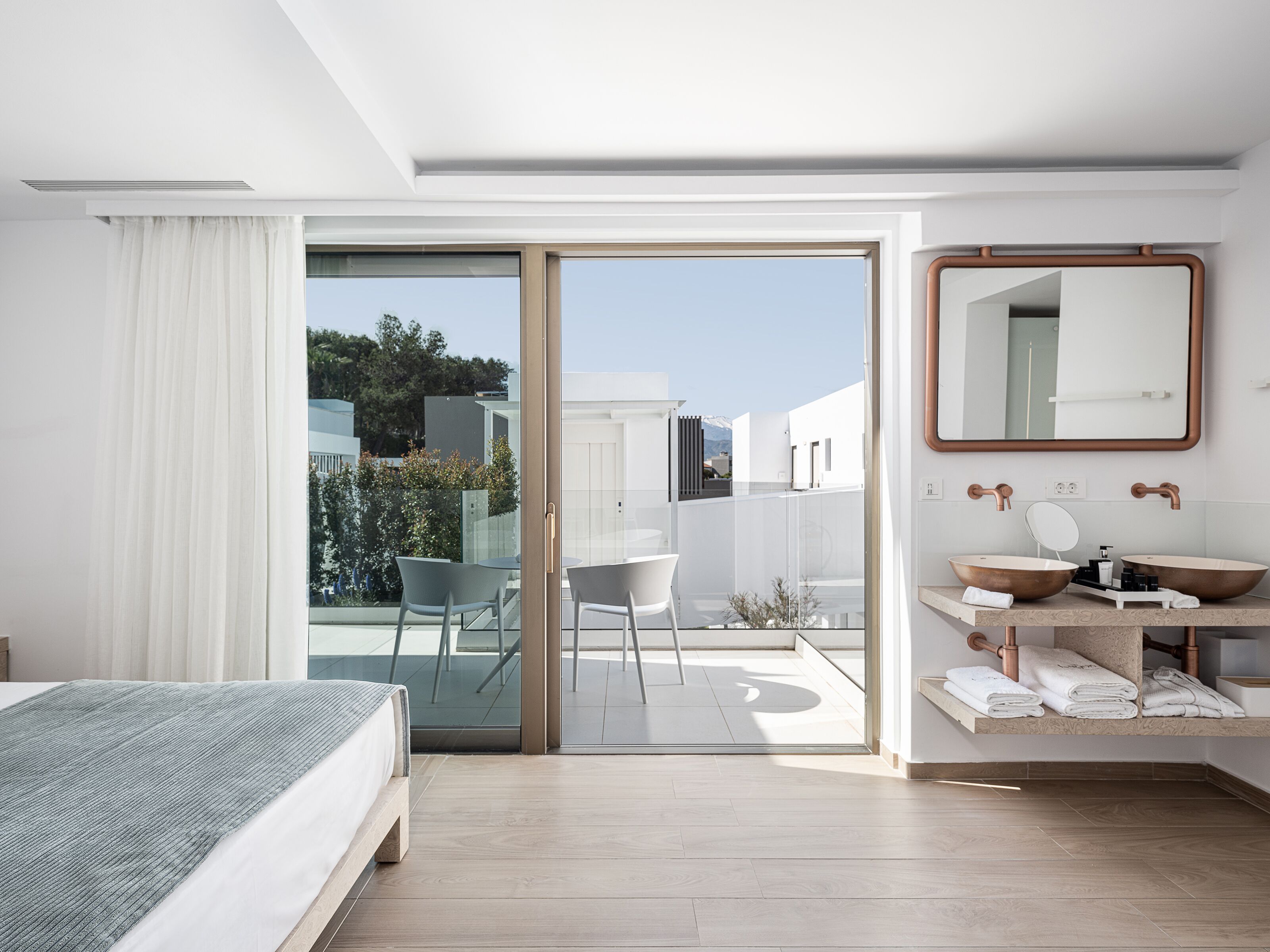 A modern hotel room with a bed, a bathroom with a large mirror, and a balcony with chairs.