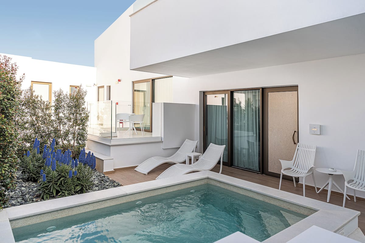 Un área de piscina exterior moderna con sillas de descanso y una pequeña mesa, rodeada por un edificio blanco con ventanas amplias y un acceso de vidrio.