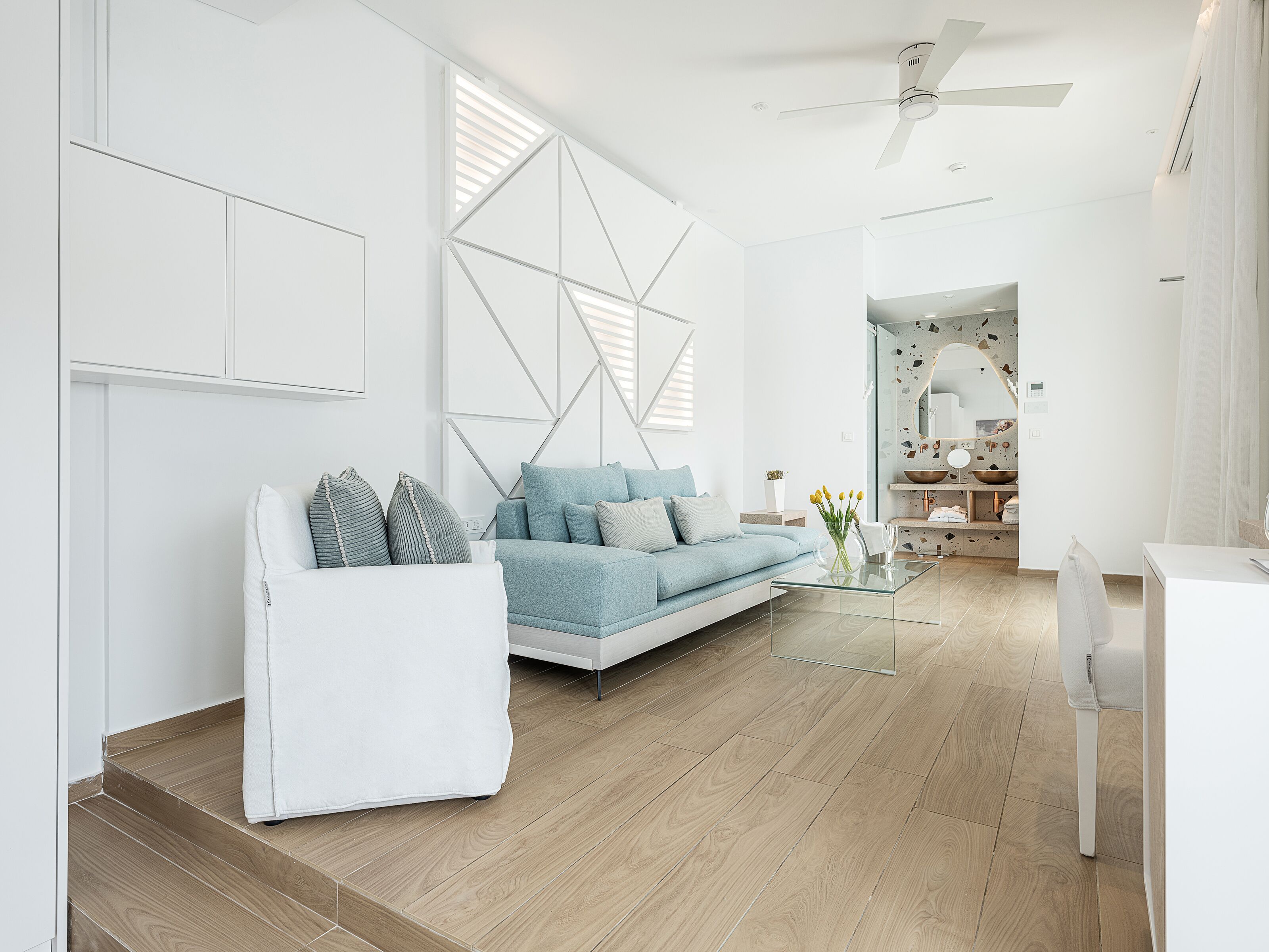 A modern living room with a light blue sofa, white armchair, and glass coffee table. A white cabinet and a geometric wall design are visible.