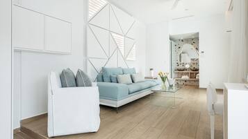 A modern living room with a light blue sofa, white armchair, and glass coffee table. A white cabinet and a geometric wall design are visible.